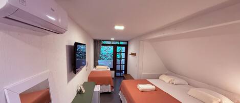 Habitación de hotel con dos camas, un escritorio, televisión y balcón con vistas a zonas verdes.