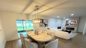 Una sala de estar moderna con comedor, que incluye una gran mesa de comedor con sillas, un sofá y un televisor de pantalla plana montado en la pared.