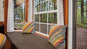 A room with a window seat, two colorful pillows, and a view of a forest and a house. - Mt Hood Cabin Sanctuary with Hot Tub, Pets Welcome (Mount Hood Village)
