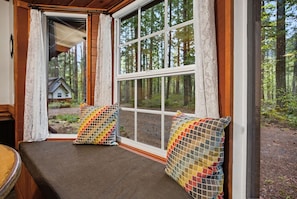 Ein Zimmer mit Fensterbank, zwei farbenfrohe Kissen und Blick auf einen Wald und ein Haus.