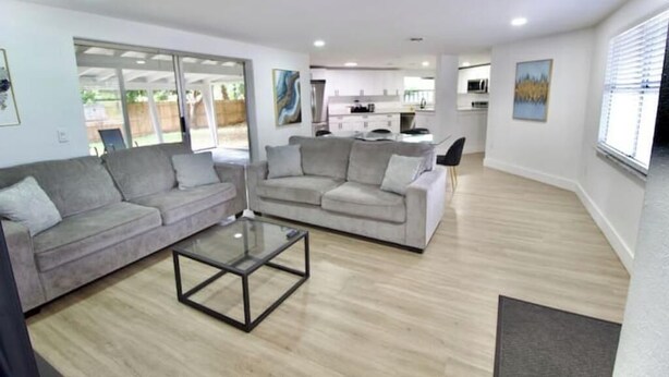 A modern living room with two sofas, a glass coffee table, and a kitchen in the background.
