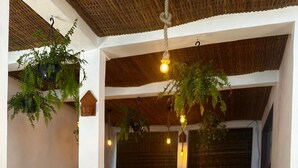 Disposição de café da manhã com diversos pratos, incluindo frutas, doces e bebidas, sobre uma mesa em um ambiente aconchegante e bem iluminado, com plantas pendentes e teto rústico.