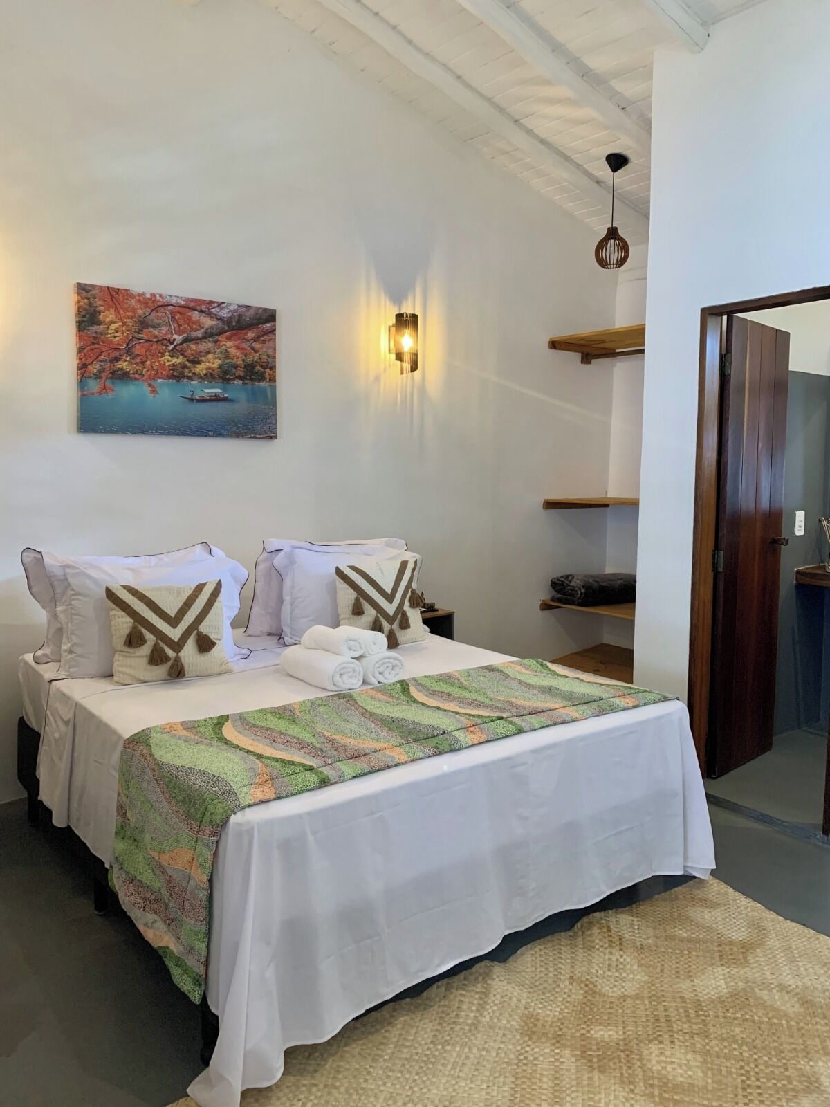 A neatly made bed with a colorful blanket, a painting of a lake with boats, and a wall-mounted light fixture.