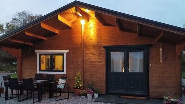 Une cabane en bois avec une porte d’entrée sombre, une fenêtre dotée d’un cadre jaune et un petit sapin de Noël.