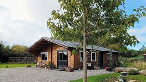 Une maison de campagne en bois, avec une allée en gravier, un porche à l’avant doté de chaises et un petit jardin.