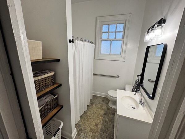 Baño pequeño con inodoro, lavamanos y ventana. Se observan estantes con cestas y una cortina de ducha.