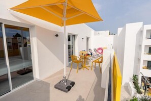Un espace extérieur moderne avec un parasol jaune, une porte vitrée donnant sur une pièce et des chaises jaunes disposées autour d’une petite table.