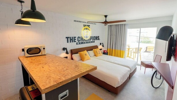 A hotel room with a bed, a desk, a microwave, a ceiling fan, and a wall with a sports team logo. - Ryans Lolas (Sant Josep de sa Talaia)