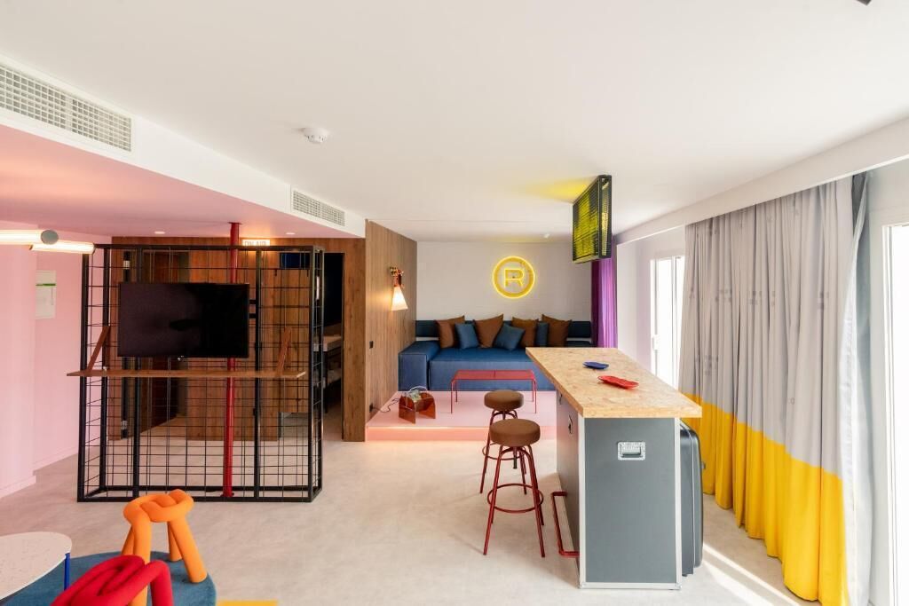 A modern hotel room with a living area, a kitchenette, a TV, and a bar area.