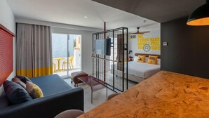A modern hotel room with a large bed, a sofa, a wooden table, and a TV. There is a balcony with a yellow table and chairs. - Ryans Lolas (Sant Josep de sa Talaia)