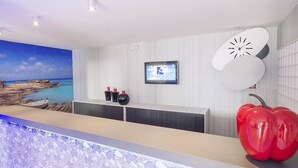 A reception area with a large painting of a beach scene, a flat-screen TV, a clock, and a decorative fruit sculpture. - Ryans Lolas (Sant Josep de sa Talaia)