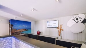 A modern reception area with a pool, a check-in/out screen, a clock, and a coastal mural. - Ryans Lolas (Sant Josep de sa Talaia)