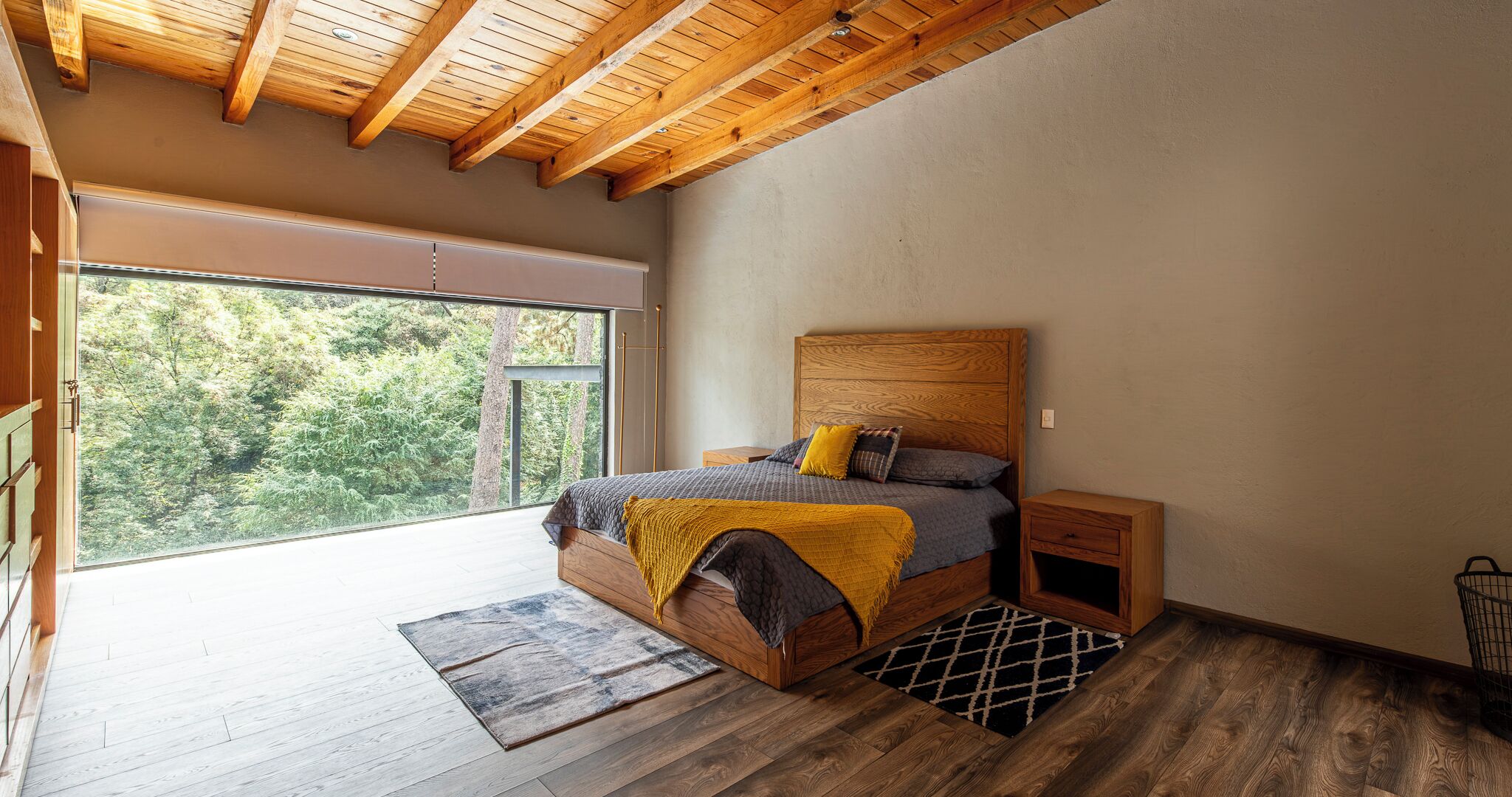 A bedroom with a large window, wooden ceiling beams, a bed with a wooden headboard, and a nightstand.