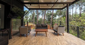 Un área de estar moderna al aire libre con cerramiento de vidrio, dos sillas de mimbre, una mesa de madera y vistas a una zona boscosa.