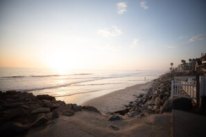 A beach with a rocky shoreline, a white fence, and buildings in the background. - Spacious 2BR - Patio + Sleeps 6 - Steps to Beach (Oceanside)