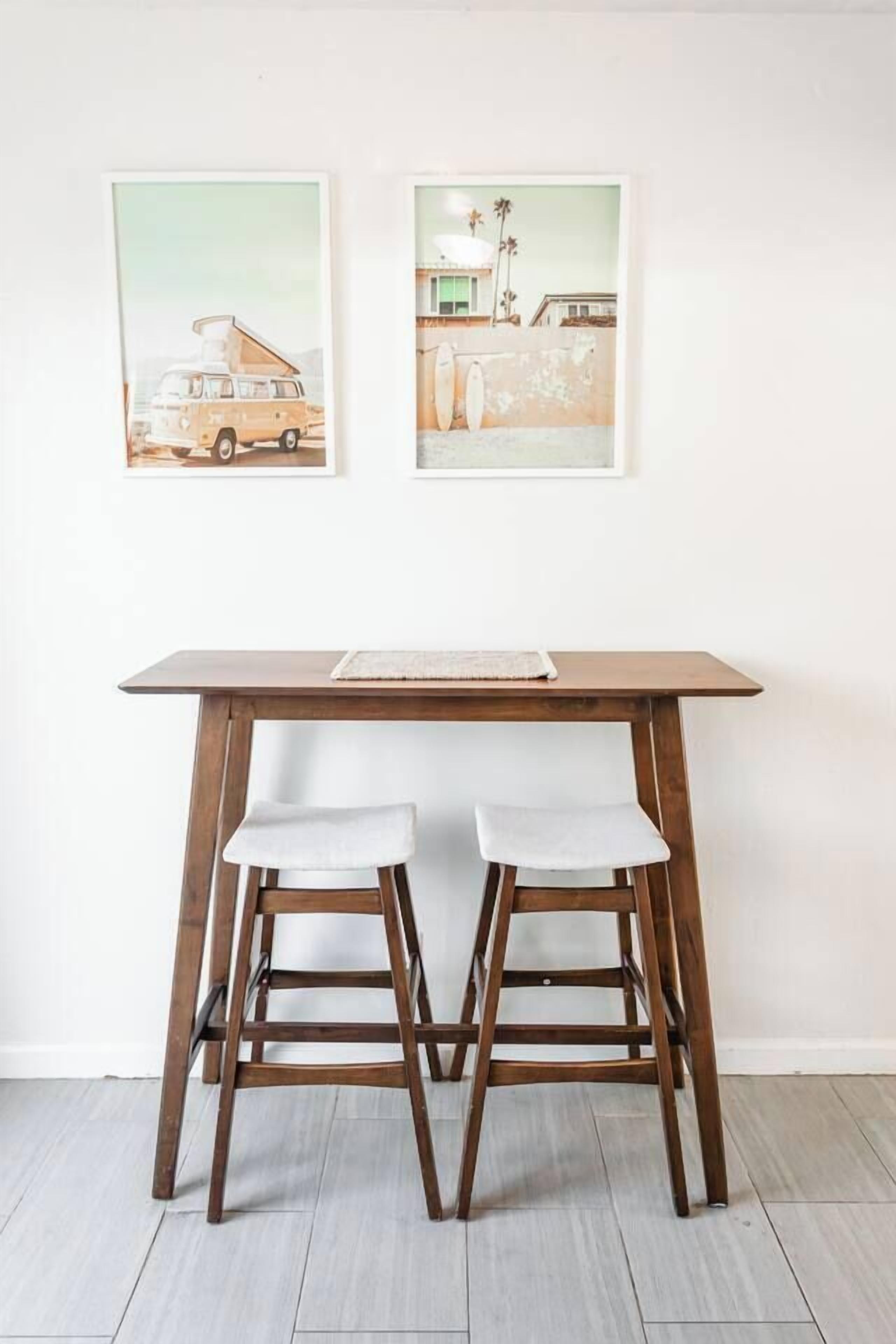 Une table en bois, deux chaises blanches avec des coussins, un tableau encadré au-dessus, et un plancher de couleur claire.