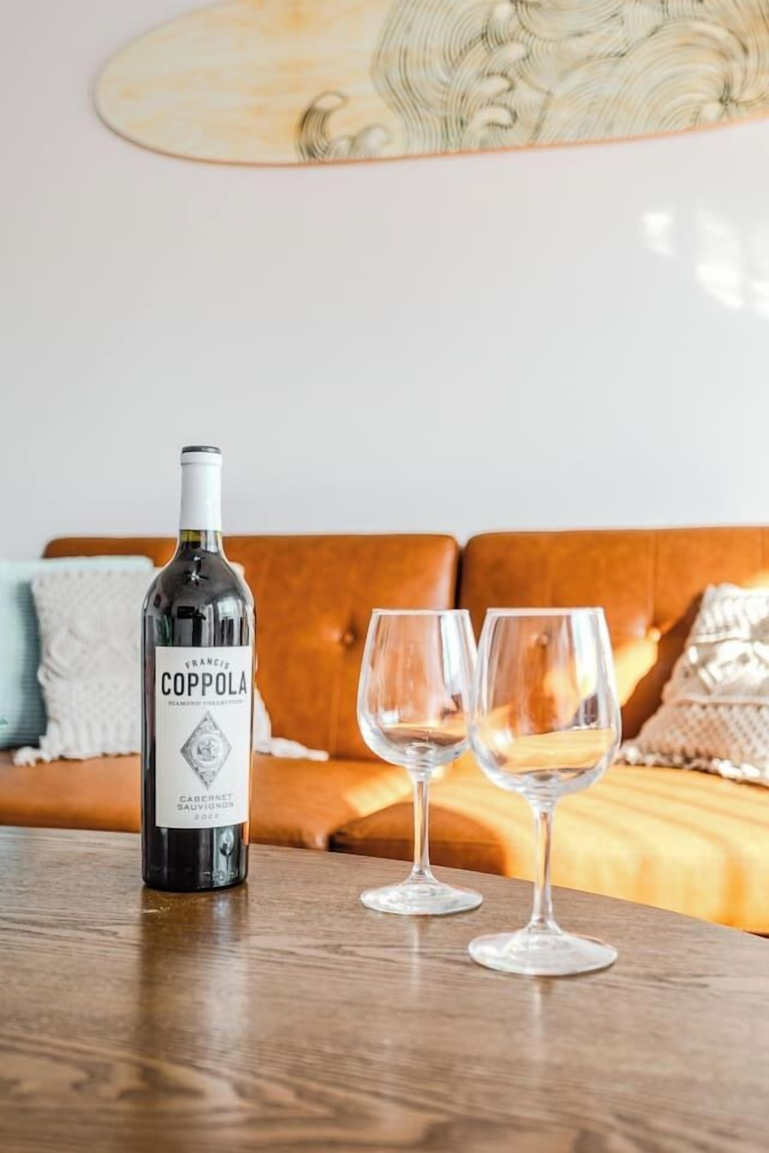 Eine Flasche Coppola-Wein und zwei Gläser auf einem Holztisch in einem Wohnzimmer mit braunem Sofa und dekorativen Kissen.
