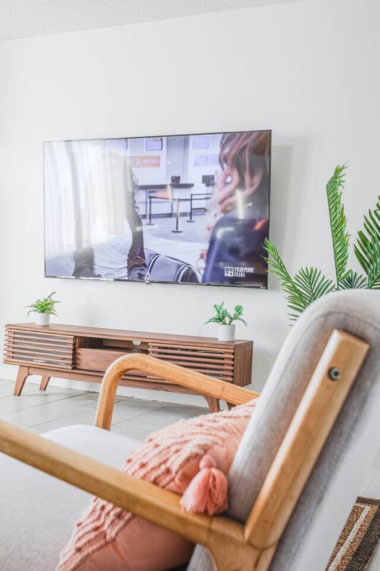 A living room with a television mounted on the wall, a wooden bench, and two potted plants.