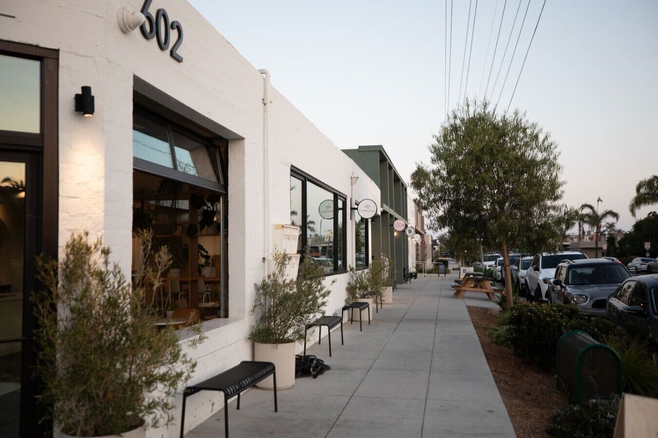 A street view with a building labeled '602' and a sidewalk lined with parked cars and potted plants.
