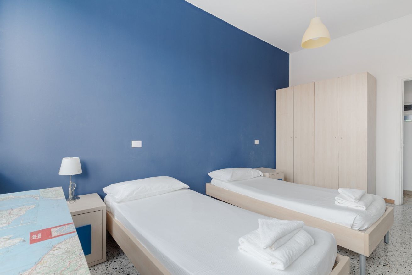 A room with two single beds, a wooden wardrobe, a map on the table, and a lamp.