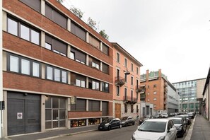 A multi-story building with a brick facade, large windows, and a parking area with cars.