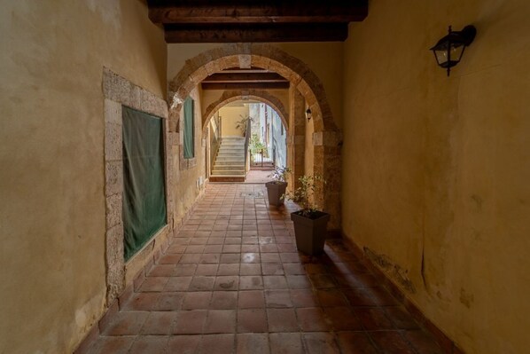 Un étroit couloir dallé de pierres, avec des portes en arc de cercle, un escalier et un pot de fleurs.