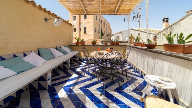Eine Dachterrasse mit gemustertem Boden, einer Sitzecke mit Kissen, einem Tisch mit Stühlen und Topfpflanzen.