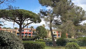 A park with trees, a building with balconies, and a clear sky. - Appt 2p-climatisé-WIFI-Parking-Pied dans l'eau-Plage La Badine-Presqu'ile Hyères (Hyères)
