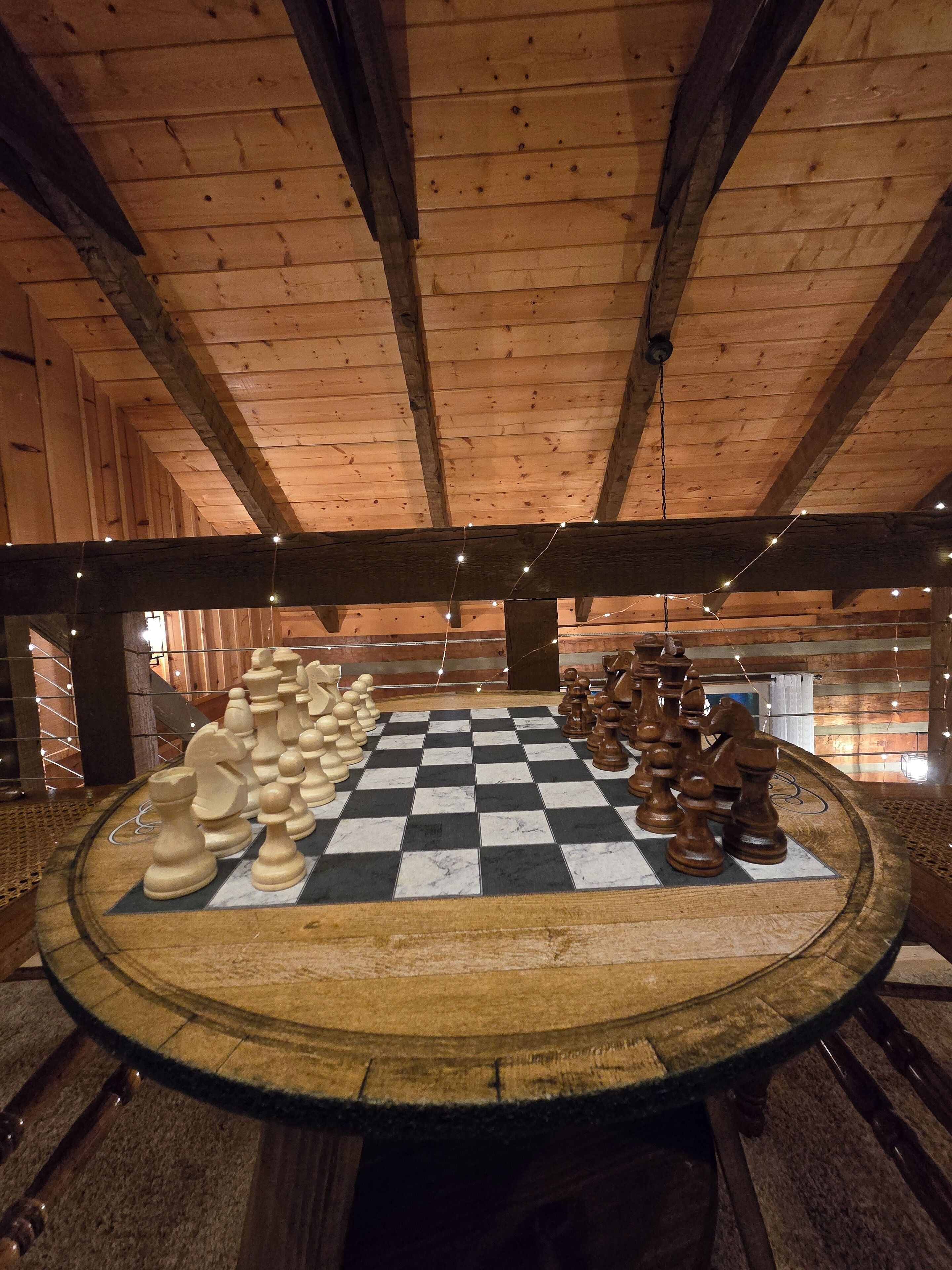 A large wooden chessboard set up in a rustic room with exposed wooden beams and string lights.
