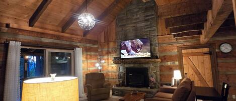 A cozy log cabin interior with a fireplace, wooden ceiling beams, a television, and comfortable seating.