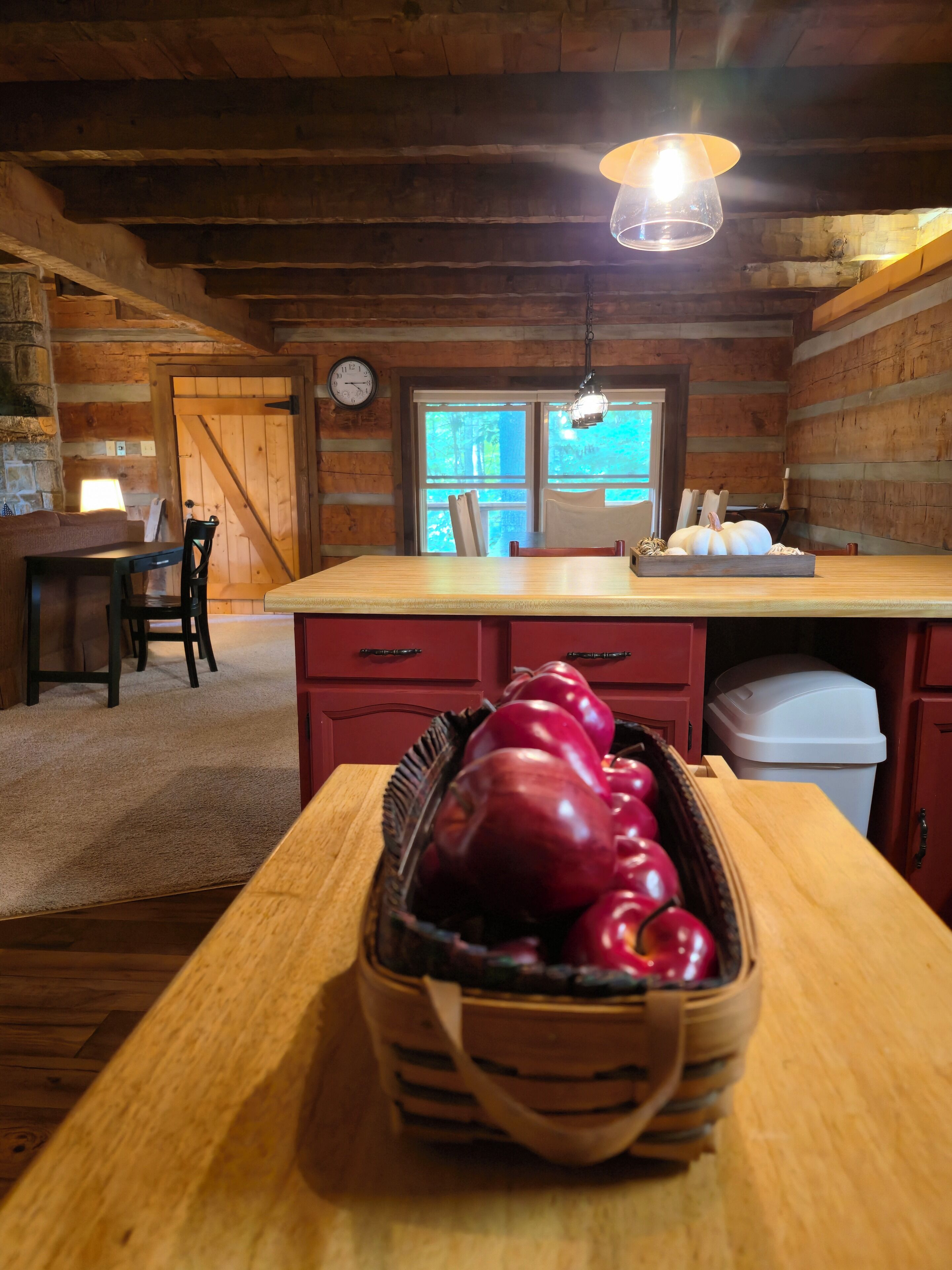 Een houten blokhut van binnen met een eethoek, een houten tafel met een mand rode appels, een houten bank en een raam met uitzicht op bomen.
