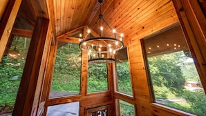 A wooden cabin interior with a central chandelier, large windows, and a view of a forested area. - Legends Lookout! Mtn Views, Bear sightings & More (Gatlinburg)