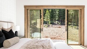 Ein Schlafzimmer mit einem großen Bett, einem Nachttisch, einer Lampe und einer Glastür, die zu einer bewaldeten Außenanlage führt.