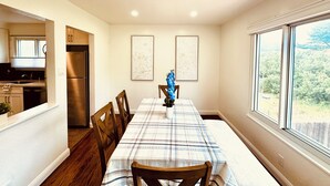 Un comedor con una mesa larga, sillas de madera y un jarrón con flores. Se ve una cocina al fondo, con un refrigerador y armarios.
