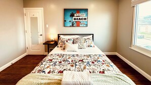 Un dormitorio con cama, una mesita de noche, una lámpara, un cuadro de playa y una ventana con vistas.