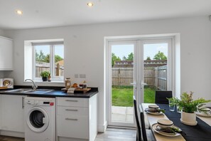 Une cuisine moderne équipée d’un lave-linge intégré, un coin repas avec une table pour deux personnes et une porte-fenêtre coulissante donnant sur un jardin.