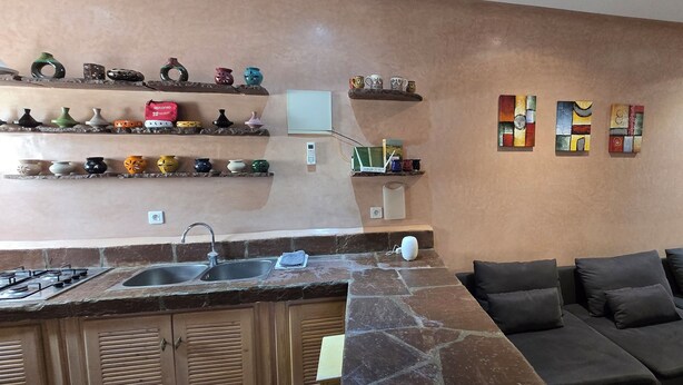Una cocina con encimera de piedra, un lavabo y una estufa. Hay estantes con adornos y un televisor montado en la pared. Un sofá con cojines se encuentra en la zona de estar.