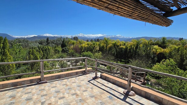 A balcony with a stone-tiled floor and metal railing, overlooking a mountainous landscape with snow-capped peaks and dense greenery.