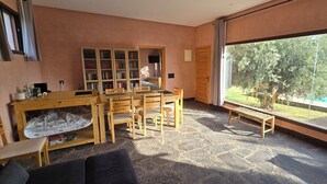 A room with a stone floor, wooden furniture, a large window with a view of a swimming pool and greenery, and a bookshelf.