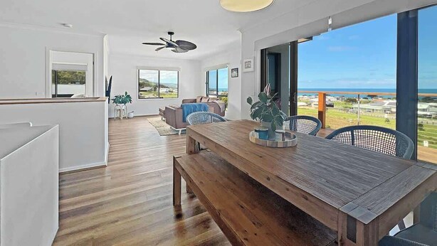 Un interior moderno con una mesa de comedor de madera, un ventilador de techo y grandes ventanas con vistas al océano.