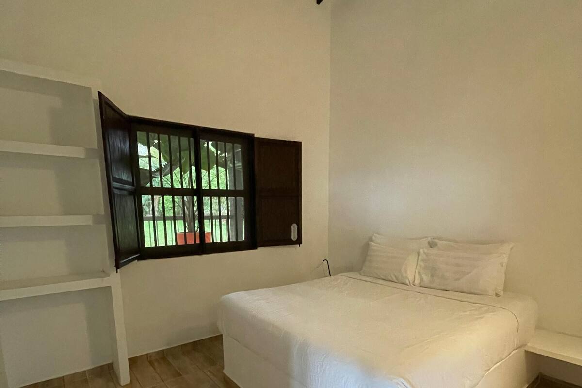 A bedroom with a bed, a window with a view of greenery, and a wooden floor.