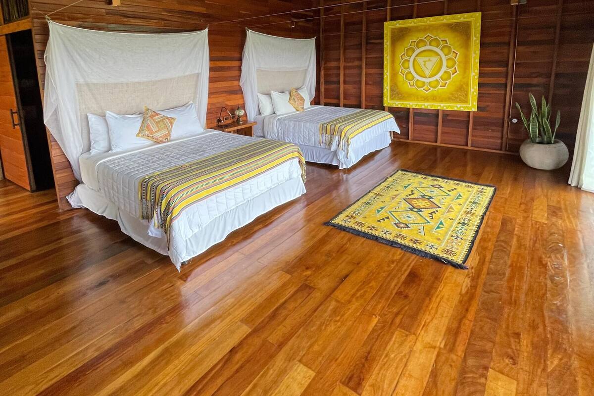 A room with two beds, a ceiling fan, a yellow wall decoration, and a wooden floor.