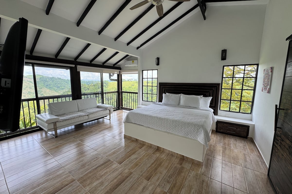 A bedroom with a large bed, a sofa, and a balcony with a view of green hills.