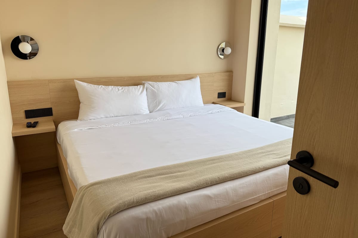 Habitación de hotel con cabecera de madera, ropa de cama blanca, mesita de noche con un reloj y una puerta con pomo.