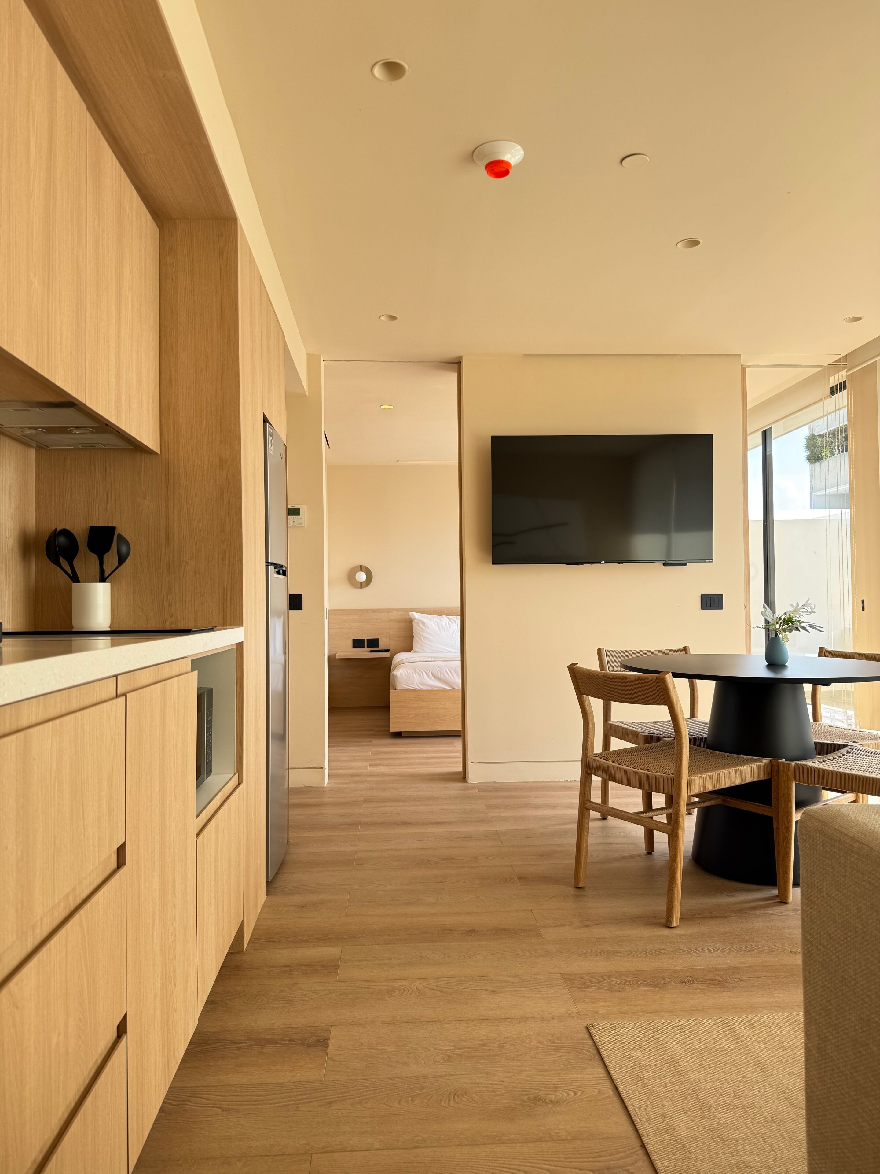 A modern kitchen with wooden cabinets, a dining area with a round table and chairs, a flat-screen TV mounted on the wall, and a bedroom visible through an open door.