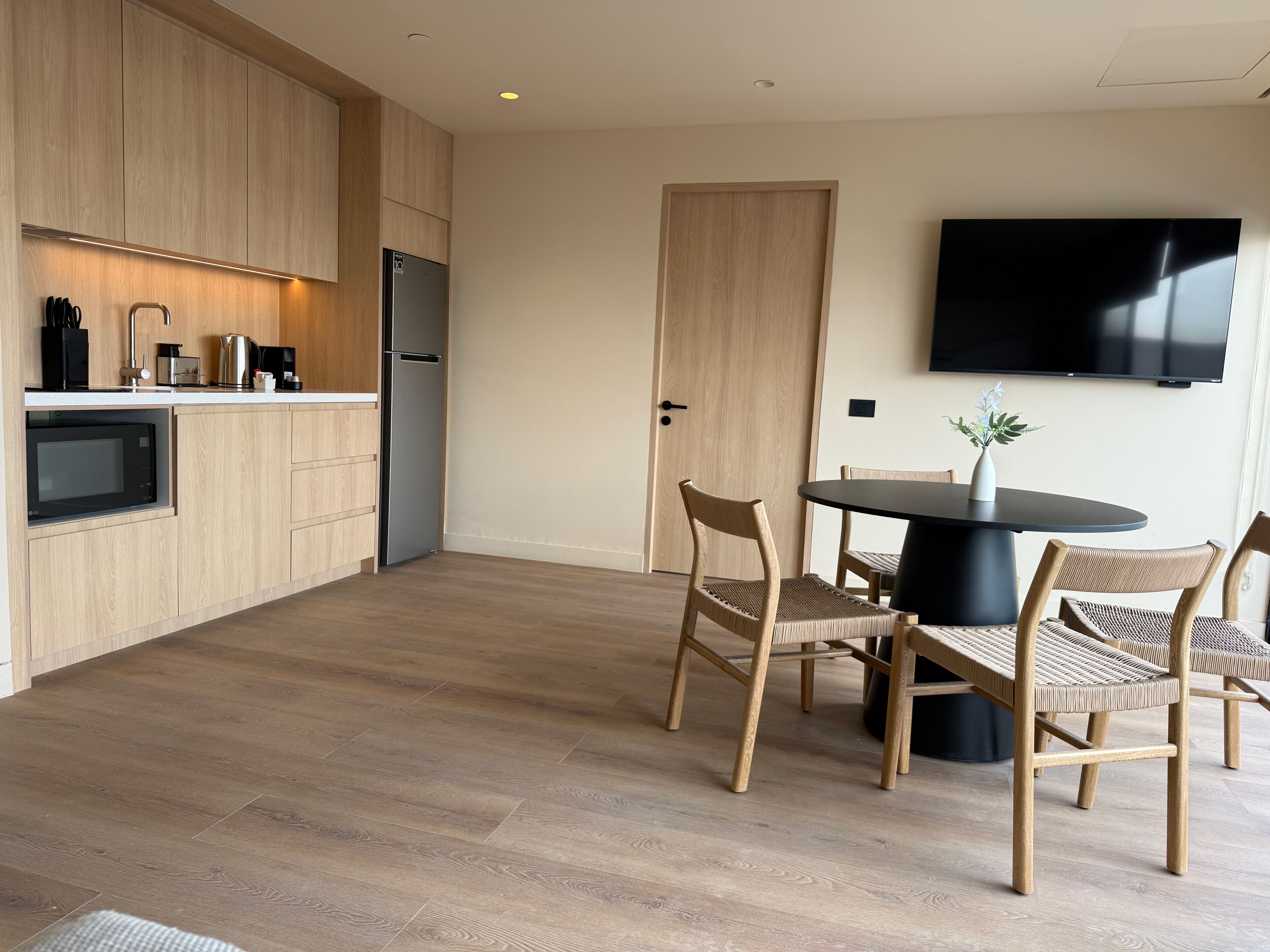 A modern kitchen with wooden cabinets, a built-in microwave, and a refrigerator. There is a dining area with a round table and four chairs. A flat-screen TV is mounted on the wall.
