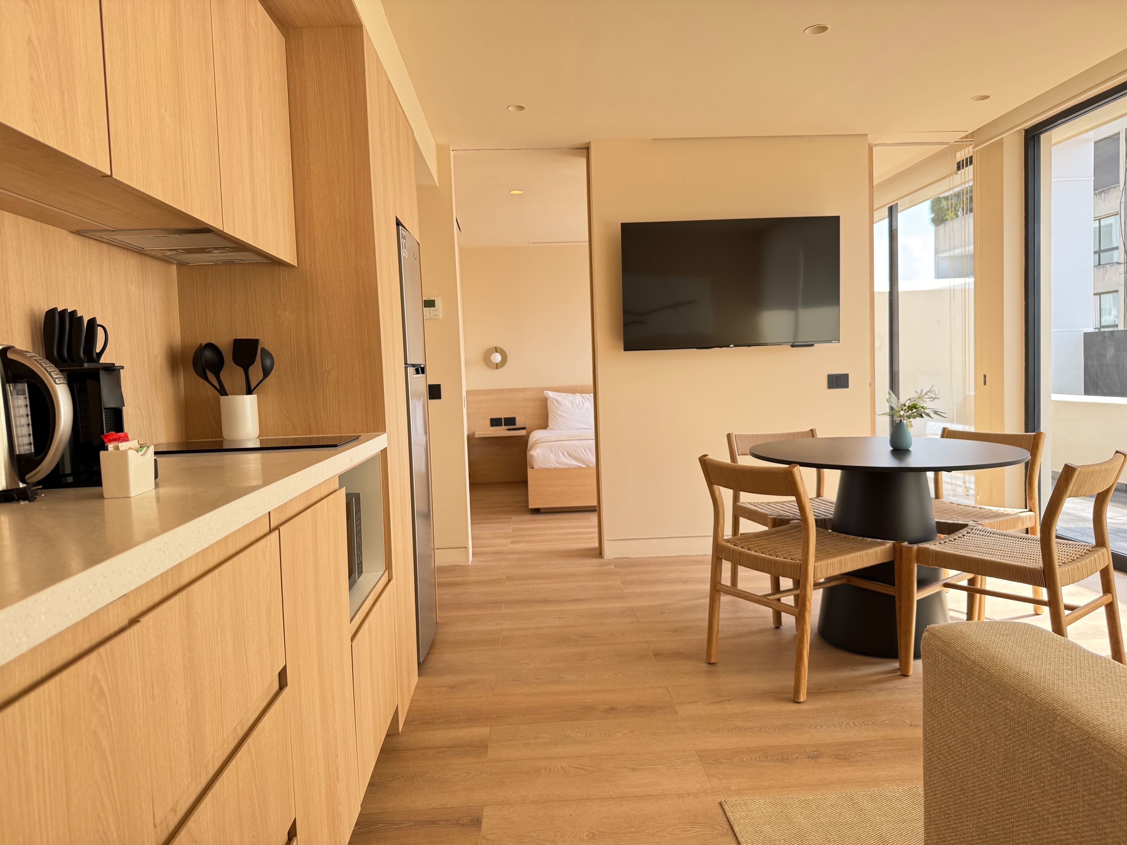 A modern kitchen with wooden cabinets, a dining area with a round table and chairs, a flat-screen TV mounted on the wall, and a bed in the adjacent room.