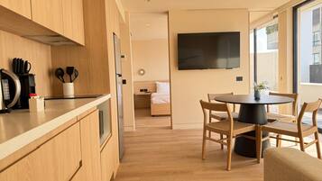 A modern kitchen with wooden cabinets, a dining area with a round table and chairs, a flat-screen TV mounted on the wall, and a bed in the adjacent room.