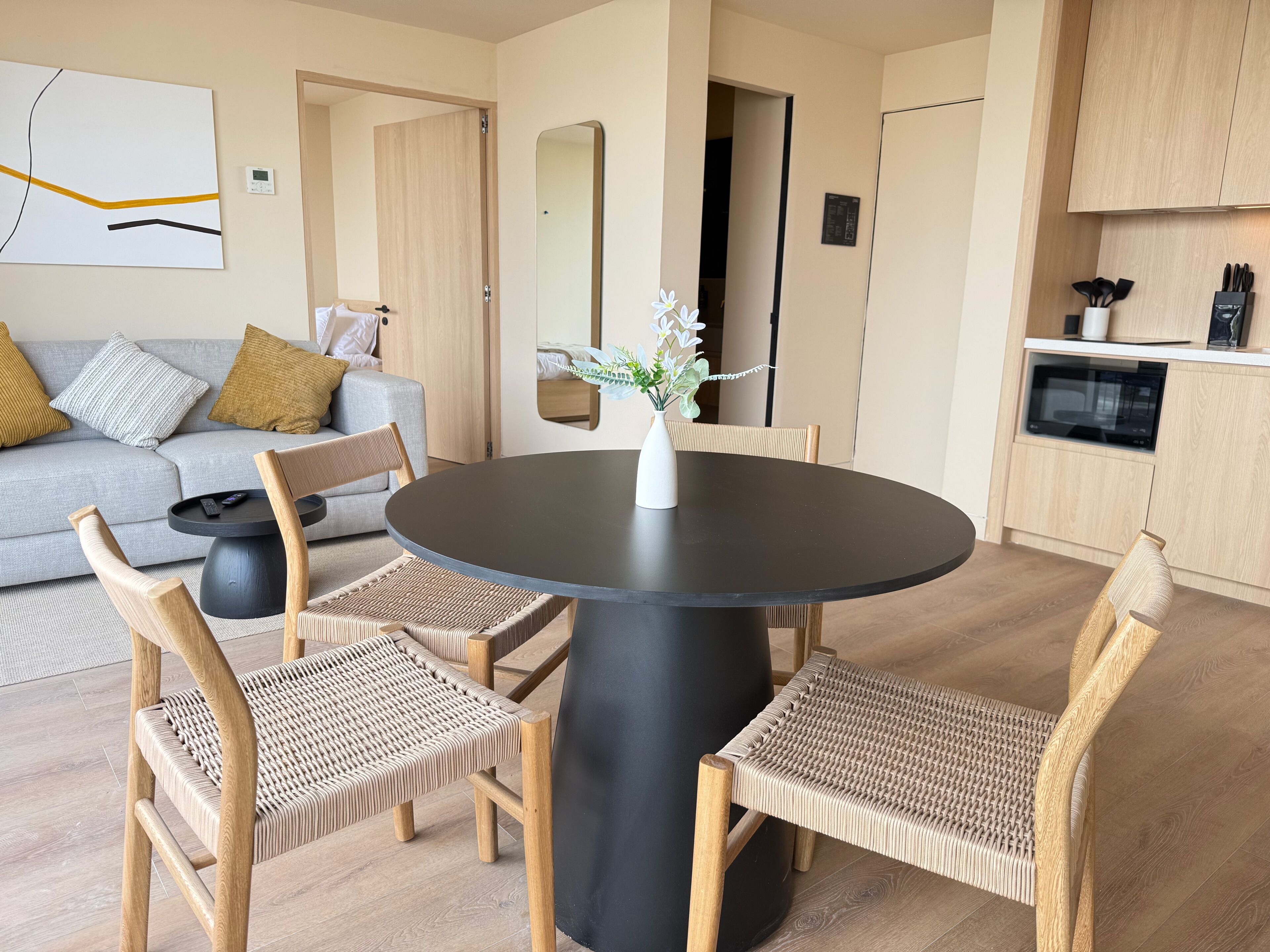 Ein modernes Wohnzimmer mit einem runden schwarzen Tisch, Korbstühlen, einem grauen Sofa mit Kissen, einer kleinen weißen Vase mit Blumen und einer Küchenzeile mit Holzschränken und Geräten.