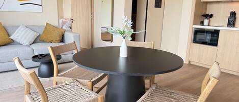 Una sala moderna con una mesa redonda negra, sillas de mimbre, un sofá gris con cojines, un jarrón blanco pequeño con flores, y una zona de cocina con gabinetes de madera y electrodomésticos.